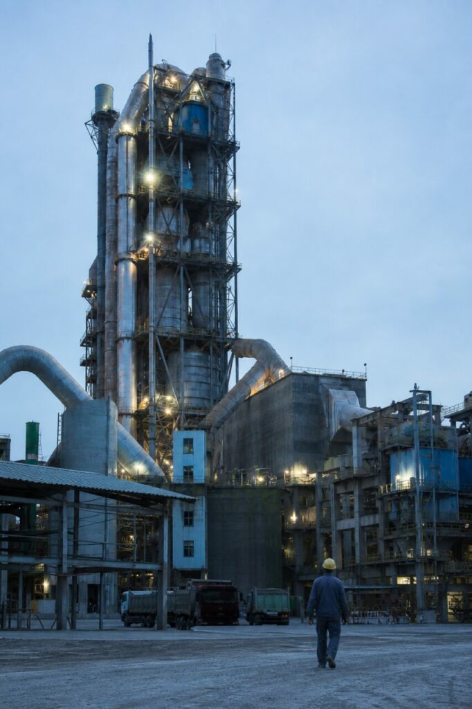Met een lange gebouwenrij langs de lage heuvels — Cementfabriek met draaitrommeloven en silo’s