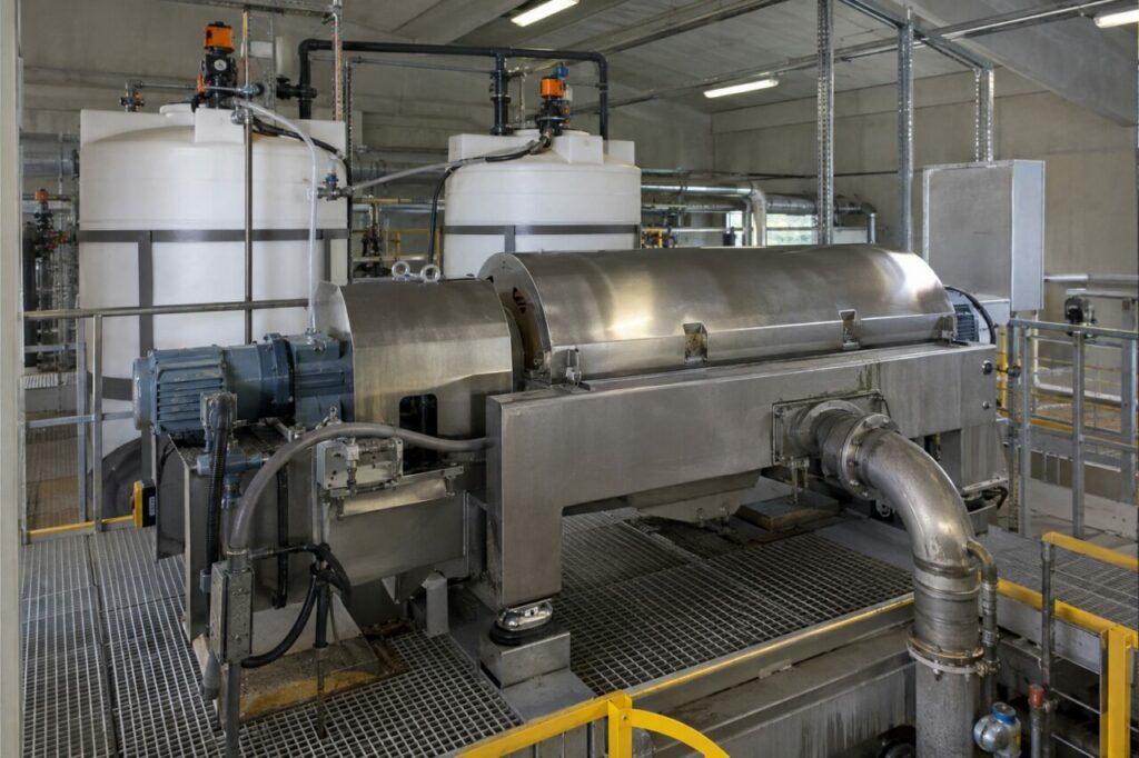 Centrifugeuse de déshydratation — une centrifugeuse de déshydratation des boues reliée à des conduites d’alimentation et des cuves de polymère, avec machines en acier inoxydable et sol caillebotis